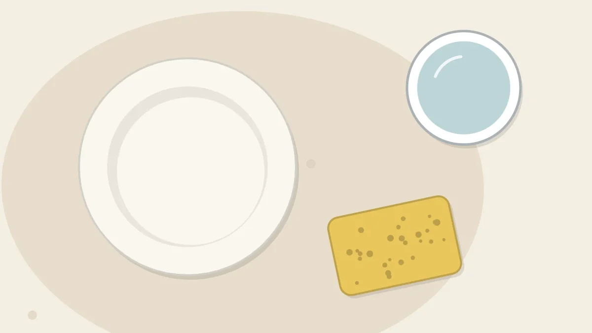 A smooth white clay dish next to a damp yellow sponge and a cup of water on an oat-colored surface